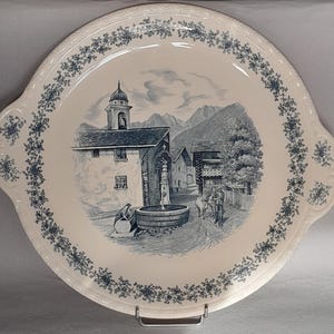 May include: A round cream-colored ceramic serving plate with two side handles, featuring a blue transferware illustration of a village scene with a fountain, a church, mountains, and a person with a cow, bordered by a delicate blue floral vine pattern.