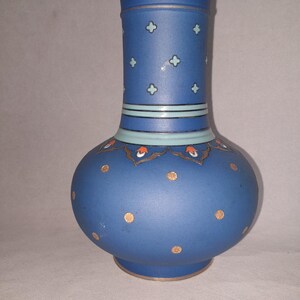 Peut inclure: Un vase en céramique bleu mat avec un corps bulbeux et un col cylindrique. Le col présente des motifs en forme de croix bleu clair, tandis que le corps est décoré de points dorés et d'une bordure festonnée avec des accents orange et blancs.