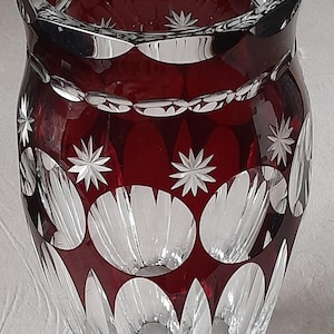 Peut inclure: Un vase en cristal taillé rouge profond avec des motifs géométriques complexes, incluant des facettes circulaires et des gravures en forme d'étoile. Le bord est festonné et la base présente un design en verre transparent à facettes.