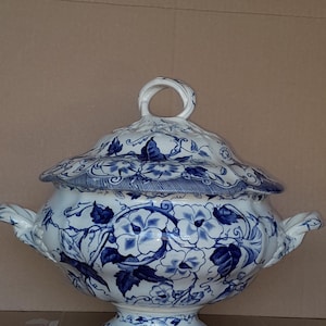 May include: A white and blue ceramic tureen with a lid, featuring a floral pattern. The tureen has two handles and a loop handle on the lid. The design includes blue flowers and leaves on a white background. The tureen is on a pedestal base.