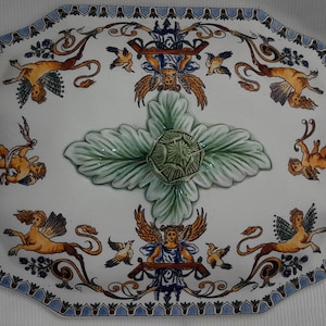 May include: An octagonal white ceramic serving platter with a detailed design. The platter features a central green leaf design and is adorned with winged figures, birds, and blue accents around the rim. The overall design is ornate and classical.