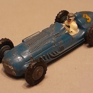 Könnte beinhalten: Ein blaues Spielzeug-Rennauto aus Metall mit einer weiß gekleideten Fahrerfigur im Cockpit, schwarzen Stollenreifen und einem gelben Aufkleber mit der Nummer drei am Heck. Das Auto hat eine lange Karosserie mit geformten Lüftungsschlitzen.