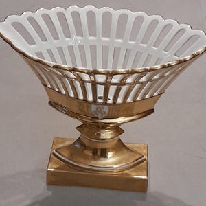 May include: A white porcelain bowl with a lattice-style rim and a gold-colored finish on the exterior base and stem. The bowl features a scalloped edge and a square pedestal base, with a small white emblem centered on the gold-toned body.