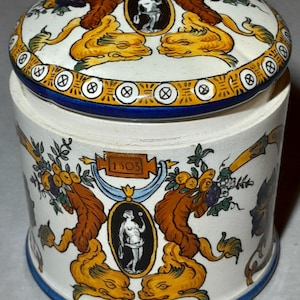 Op de afbeelding: Een cilindrische keramische pot met bijpassend deksel, voorzien van een crèmekleurige basis met handgeschilderde motieven. Het ontwerp bevat oranje krulwerk, blauwe accenten en zwarte ovale medaillons met een figuur. De tekst 1503 staat op een banier.