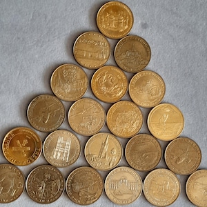 Pode incluir: Uma coleção de vinte moedas comemorativas em tom dourado dispostas em formação triangular. Cada moeda apresenta imagens em relevo únicas, incluindo marcos arquitetónicos, veículos históricos e vários símbolos culturais franceses com texto.