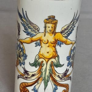 Peut inclure: Un vase cylindrique en céramique orné d'une figure ailée peinte à la main dans des tons jaune, bleu et vert. Le motif comprend des volutes décoratives et une bordure avec des cercles jaunes et noirs sur les bords supérieur et inférieur.
