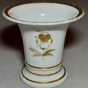 Pode incluir: Um vaso de porcelana branca com uma borda alargada e acabamento em tom dourado. O corpo do vaso é decorado com uma ilustração floral dourada e uma faixa dourada perto da base, que inclui também um delicado padrão de folhas.