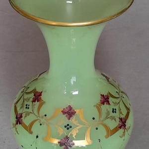 May include: A pale green glass vase with a flared rim featuring a gold-painted edge. The body is decorated with hand-painted floral motifs in dark pink and purple, accented by gold scrollwork and small dark green dots arranged in patterns.