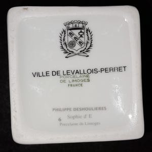 May include: A white porcelain item with text that reads "VILLE DE LEVALLOIS-PERRET PORCELAINE DE LIMOGES FRANCE". The item also features a crest and the text "PHILIPPE DESHOULIERES Sophie d' E Porcelaine de Limoges".