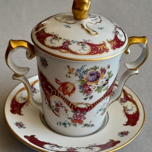 May include: A white porcelain lidded cup with two handles and a matching saucer, featuring intricate floral patterns in purple, orange, and green, accented by deep red scrollwork and gold trim, topped with a gold-coloured finial on the lid.
