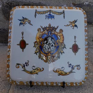 May include: Square, white ceramic tray with a decorative design. The tray features a central crest with cherubs, a crown, and a blue and gold border. The border is adorned with yellow and gold floral patterns, and the tray has a black metal stand.