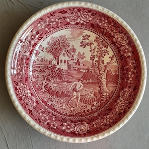 Peut inclure: Une assiette ronde en céramique ornée d'un motif de transfert rouge foncé et blanc. Le bord présente une bordure florale détaillée, tandis que le centre représente un paysage rural avec une maison, des arbres et une silhouette.
