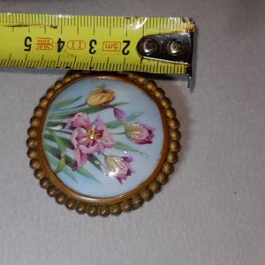 May include: A round, antique-style brooch with a floral design. The brooch features a light blue background with a painting of pink and purple flowers. The brooch is approximately 2 inches in diameter and has a gold-colored, beaded edge.