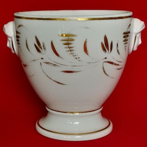 May include: A white porcelain vessel featuring a gold-painted leaf and vine pattern around the exterior. The container has a flared rim, a pedestal base, and two white sculpted handles on the sides, each shaped like a human face in profile.