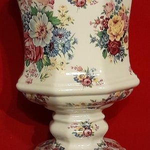 May include: A cream-colored ceramic vase with a hexagonal shape and a pedestal base. The surface features a colorful floral pattern with roses and small blue flowers. The rim and base edges are accented with a thin gold-colored trim.