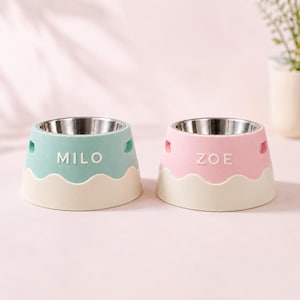 Pode incluir: Dois recipientes para animais com inserções de aço inoxidável. Um é azul-claro com o nome MILO em letras brancas e o outro é rosa-claro com o nome ZOE em letras brancas. Ambos possuem uma base ondulada em tom creme e aberturas laterais.