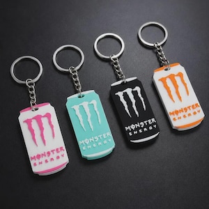 Pode incluir: Quatro porta-chaves de metal com pendentes retangulares em forma de latas de bebida energética. Cada pendente apresenta o logótipo e o texto Monster Energy em cores diferentes: rosa sobre branco, branco sobre azul claro, branco sobre preto e laranja sobre branco.
