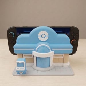 Op de afbeelding: Een lichtblauwe en beige spelconsolehouder in de vorm van een gebouw met een wit cirkelvormig embleem aan de voorzijde. De houder is voorzien van een kleine blauwe kiosk aan de linkerkant en houdt een zwarte handheld spelconsole met controllers.