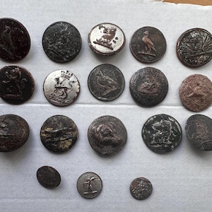 Könnte beinhalten: Eine Sammlung antiker Metallknöpfe in verschiedenen Größen und Designs. Die Knöpfe zeigen geprägte Bilder von Vögeln, Löwen und anderen Figuren. Die Knöpfe sind in Braun-, Silber- und Bronzetönen gehalten und weisen Gebrauchsspuren auf.