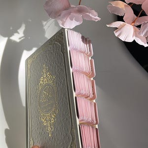 Può includere: Un libro color crema con una texture in rilievo e calligrafia araba in lamina d'oro sulla copertina. Il libro presenta numerose linguette indice rosa chiaro sul lato e le pagine hanno bordi dorati. Il design include intricati bordi dorati.