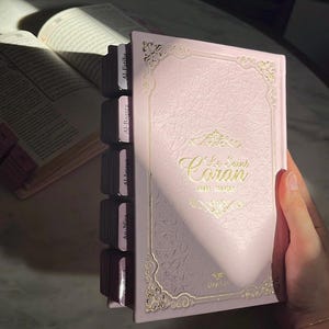 May include: A light pink book titled Le Saint Coran with gold embossed filigree borders and lettering on the cover. The book features dark pink index tabs along the side labeled with chapter names, and a second open book is visible in the background.