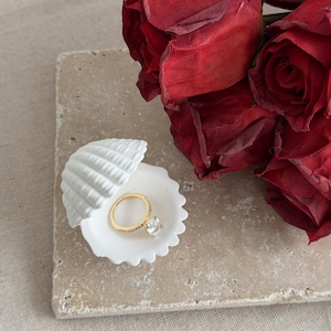 May include: A gold ring with a large, clear gemstone sits in a white seashell-shaped dish. The dish rests on a rectangular, light-colored stone surface. A bouquet of deep red roses is in the background, creating a romantic setting.