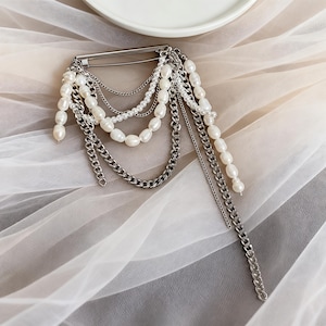 May include: A silver-tone brooch featuring multiple strands of white pearls and silver chains. The brooch has a safety pin style clasp and the pearl strands vary in length. The jewelry is displayed on a white, sheer fabric.