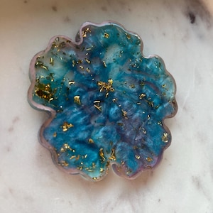 May include: A four-leaf clover-shaped resin coaster with a blue and purple marbled design. Gold leaf flakes are embedded throughout the coaster. The coaster has a light pink border.