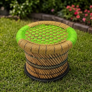May include: A round, woven stool with a green and tan colour scheme. The seat is a woven green pattern, surrounded by a tan border. The sides are made of bamboo and the base is black. The stool is sitting on green grass.