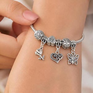 Op de afbeelding: Zilveren bedelarmband met decoratieve bedels. De armband heeft bedels in de vorm van een roos, een hart en een vlinder. De armband wordt om de pols gedragen.