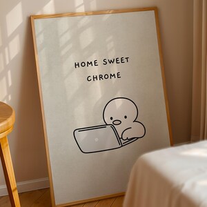 May include: A rectangular framed wall art print featuring a light beige background with the black text "HOME SWEET CHROME" above a simple black line drawing of a round-headed character sitting and working on a laptop computer.