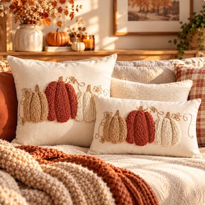 Pode incluir: Duas almofadas decorativas em tom off-white com bordado texturizado de três abóboras em tons de bege, laranja queimado e creme. As abóboras estão ligadas por ramos e pequenas flores bordadas sobre a capa de tecido liso.