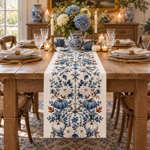 Peut inclure: Un chemin de table rectangulaire orné d'un motif répétitif de citrouilles bleues et blanches, de chrysanthèmes blancs et de feuillage bleu sur fond crème. Le chemin est centré sur une table à manger en bois avec assiettes, verres et bougies.
