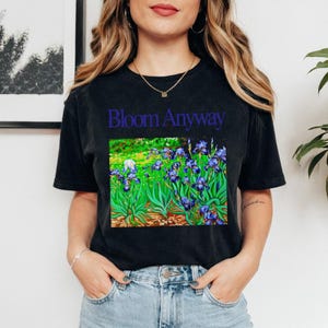 May include: Black t-shirt with the text "Bloom Anyway" above a colorful floral design featuring blue irises and green foliage. The shirt is a comfortable, casual style.