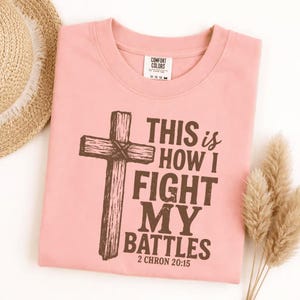 May include: A light pink t-shirt featuring a dark brown graphic of a wooden cross and the text "THIS is HOW I FIGHT MY BATTLES 2 CHRON 20:15". The shirt has a crew neckline and a Comfort Colors brand label visible on the inside of the collar.