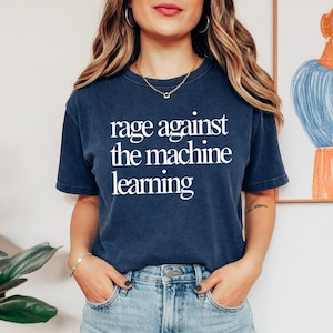 May include: Navy blue t-shirt with the white text "rage against the machine learning". The shirt is worn with blue jeans. A gold necklace and bracelet are also visible.