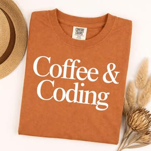 May include: A rust-colored crew neck t-shirt featuring the white text Coffee & Coding printed on the front. The shirt has a Comfort Colors brand label inside the collar. The garment is displayed alongside a straw hat and dried floral stems.