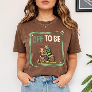 May include: Brown t-shirt with a green and white graphic that reads "OFF TO BE." The graphic features a frog riding a bicycle with a cat in the basket. The words "Radical Left Scrum" are below the main text.