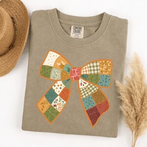 May include: A Comfort Colors olive green sweatshirt with a large, colorful bow design. The bow is made of patchwork squares in various patterns and colors, including floral, plaid, and solid hues. The bow has an orange outline.