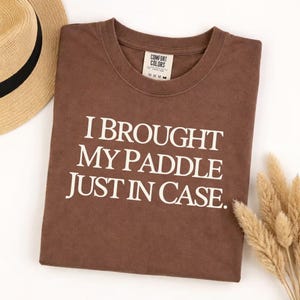 May include: A brown t-shirt with the text "I BROUGHT MY PADDLE JUST IN CASE" in white. The shirt is folded and next to a straw hat and a bundle of dried wheat.