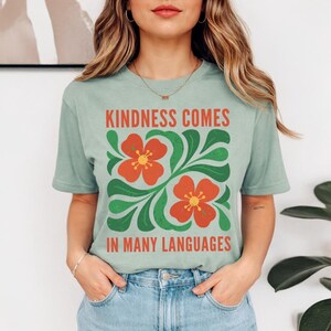 May include: A light green t-shirt with the text "KINDNESS COMES IN MANY LANGUAGES" in red and green. The shirt features a floral design with orange flowers and green leaves.