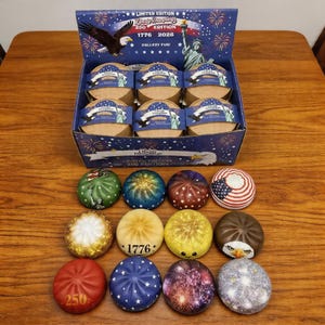 Puede incluir: Una caja expositora de Liberty Dumplings, con temática patriótica. La caja es azul marino con un águila y la Estatua de la Libertad. Las albóndigas son redondas, con varios diseños, incluyendo la bandera americana, fuegos artificiales y el número "1776".