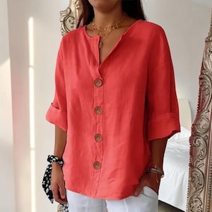 May include: A coral-colored button-down shirt with a V-neckline and three-quarter length sleeves. The shirt features four large, round, dark brown buttons down the center front and is paired with white pants. The fabric has a visible texture.