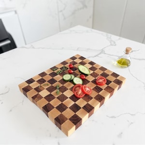 Puede incluir: Una tabla de cortar cuadrada de madera con un patrón de tablero de ajedrez de cuadrados alternos de madera de arce clara y madera de nogal oscura. La tabla está cubierta con rodajas de pepino, tomates cherry y ramitas de hierbas frescas.