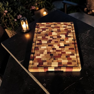 Puede incluir: Una tabla de cortar rectangular de madera con un patrón de mosaico de grano final compuesto por pequeños bloques multicolores en tonos beige claro, marrón medio y morado rojizo intenso, con un canal para jugos a lo largo del borde.