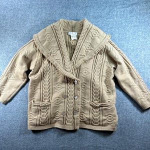 May include: A tan double-breasted cardigan featuring a wide shawl collar with fringe trim, vertical cable knit patterns, and geometric diamond motifs. The garment includes four buttons on the front, two lower pockets, and a white interior label.
