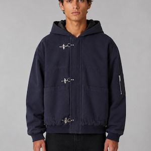 May include: A navy blue hooded jacket featuring three silver-toned metal firemen-style clasps down the front closure, two large patch pockets, a ribbed hem, and a small zippered utility pocket on the left sleeve, worn over black pants.