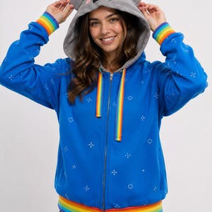 May include: A bright blue zip-up hoodie featuring a gray hood with cat ears and a pixelated face. The garment includes rainbow-striped cuffs, a rainbow-striped hem, and rainbow-striped drawstring cords. The blue fabric has a white cross-stitch pattern.