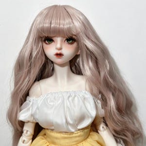 May include: A detailed close-up of a doll with long, wavy, light pink hair. The doll has a fair complexion, green eyes, and a delicate face. It wears a white off-the-shoulder top and a yellow skirt. The doll is a collectible item.