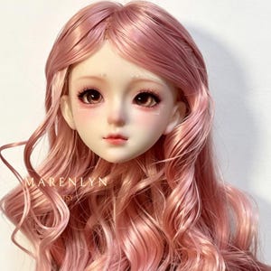 May include: A doll head with a pale complexion, rosy cheeks, and large brown eyes. The doll has long, wavy pink hair and delicate facial features. The text "MARENLYN" and "ETSY" is visible on the bottom left.
